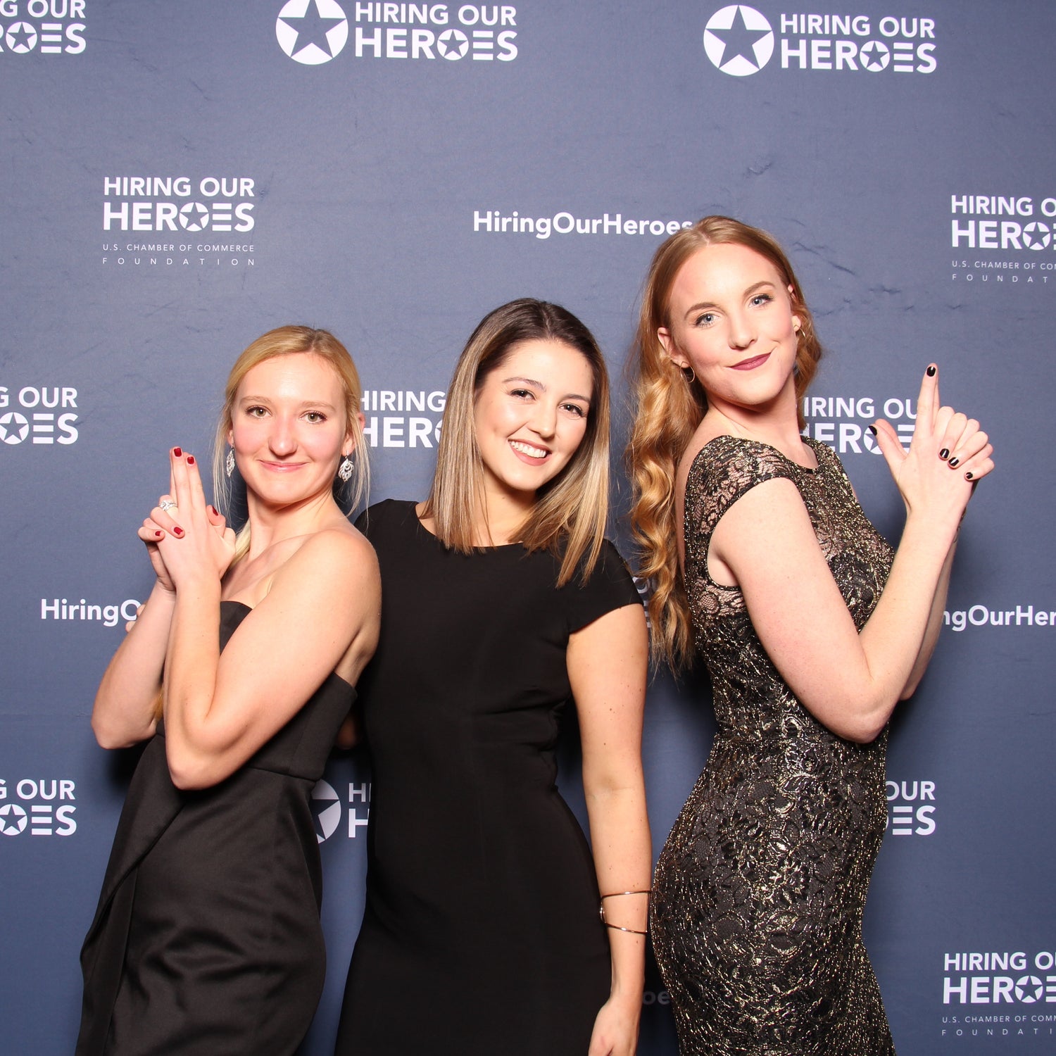 Three women posing in front of a step and repeat photo banner at a Hiring Our Heroes event