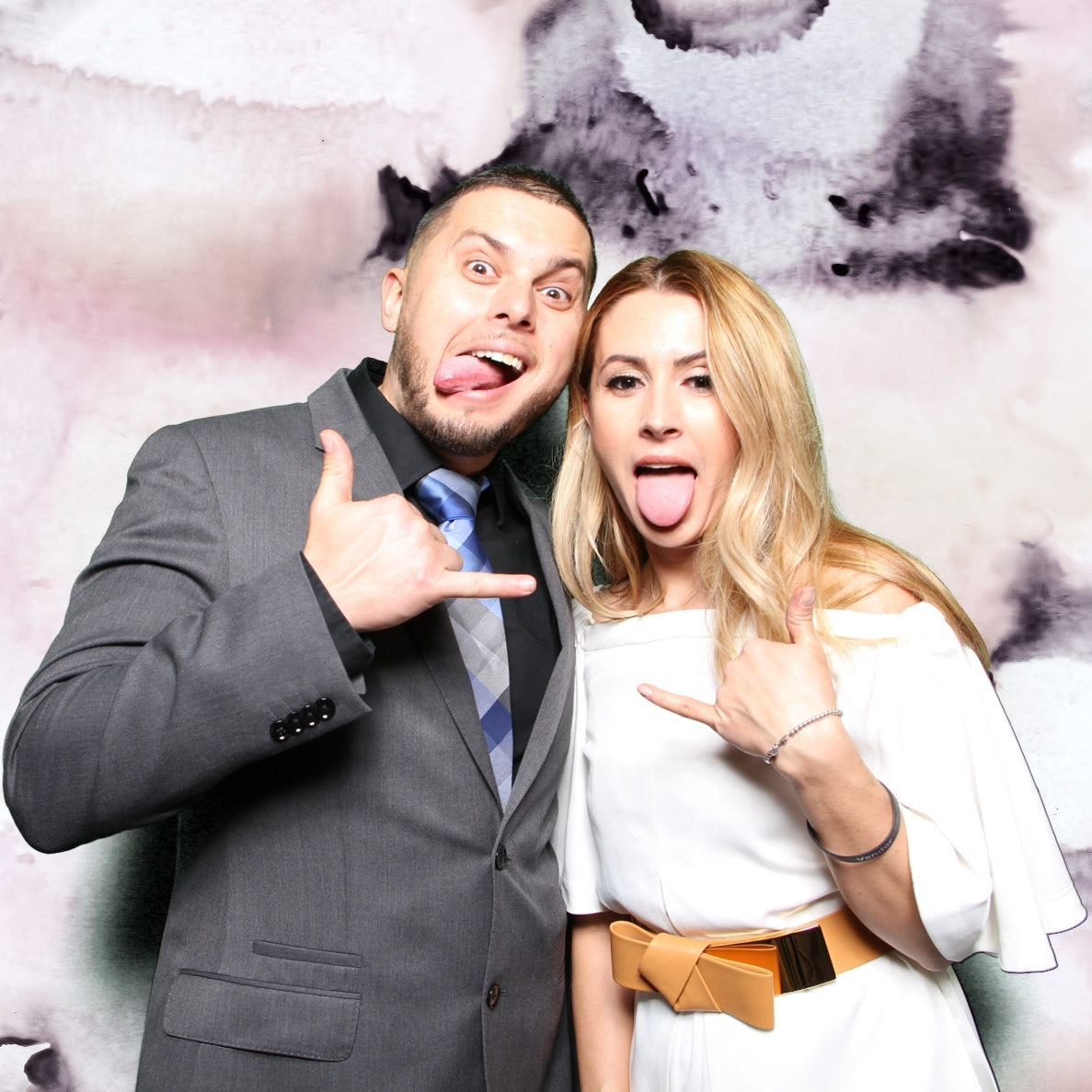Man in grey suit and woman in white dress with belt posting for photo sticking out their tongue and both holding up one hand