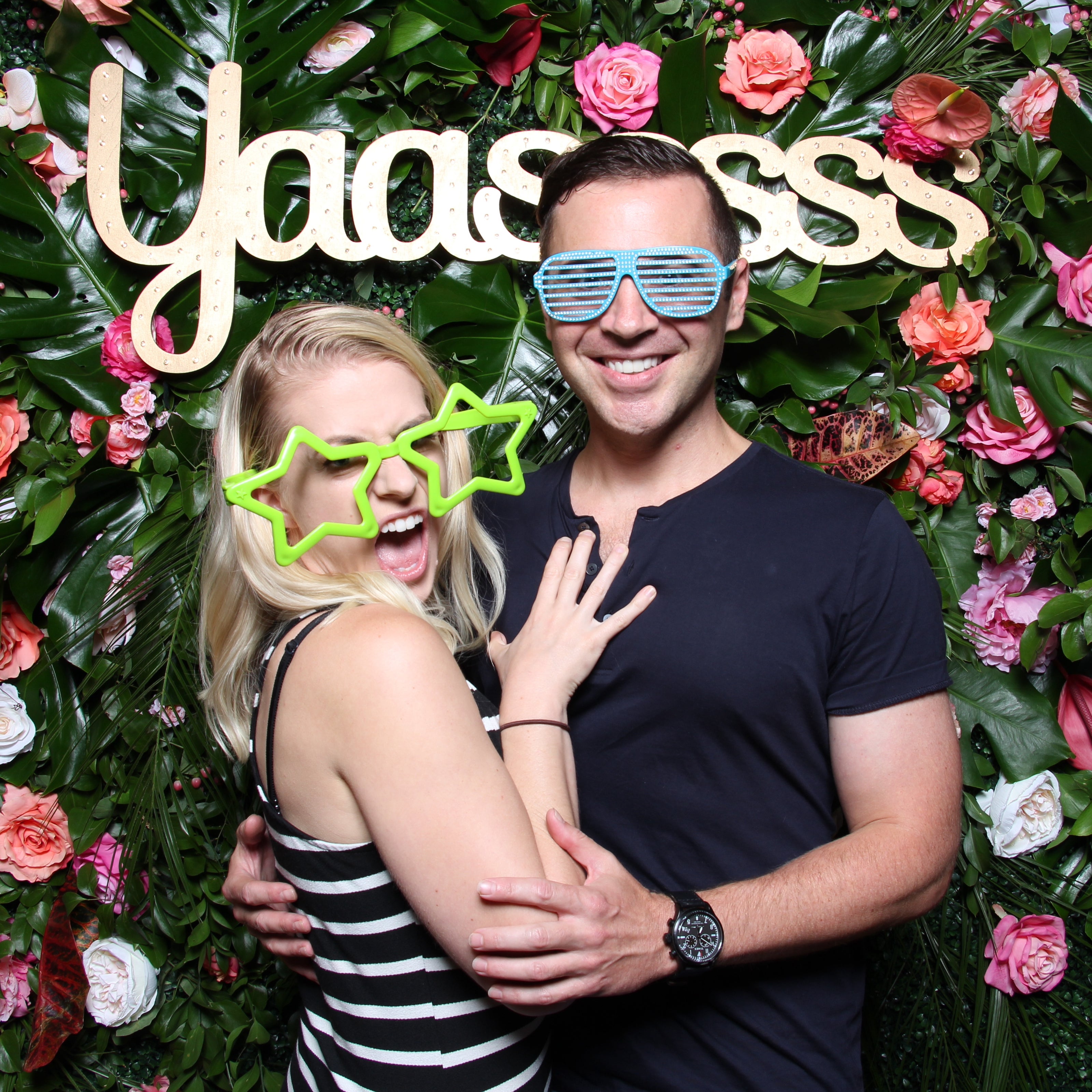 Guy and girl in a photo booth wearing crazy glasses while posing in front of a flower backdrop with a gold "Yaasssss" sign