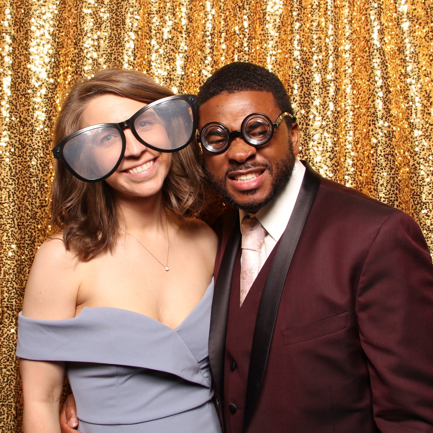 Couple in a photo booth posing with prop glasses with a sparkly gold backdrop